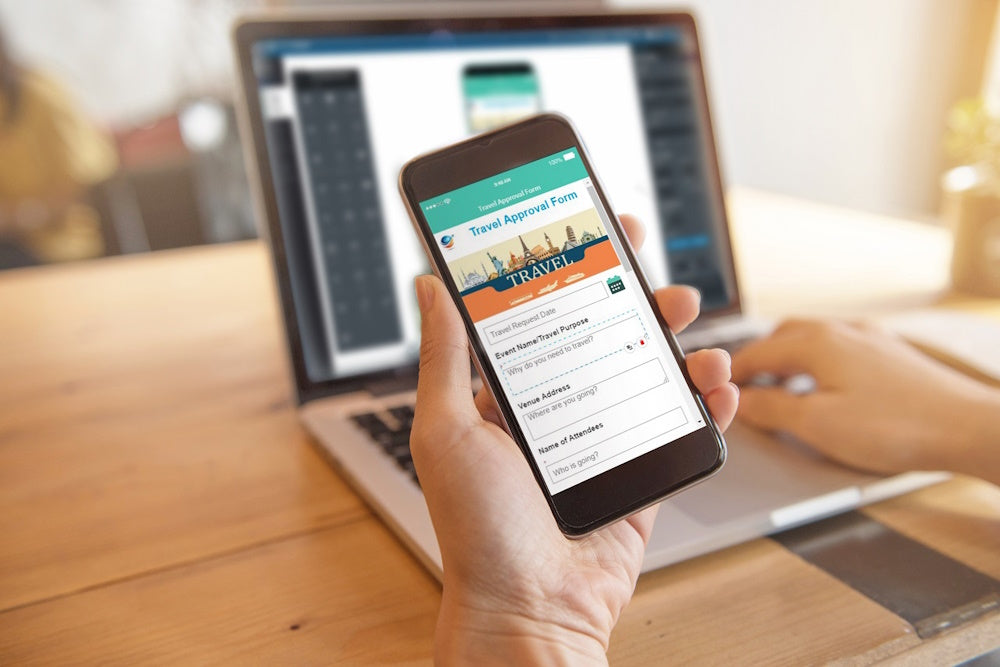 Hand håller en mobiltelefon med reseformulär, laptop i bakgrunden.
