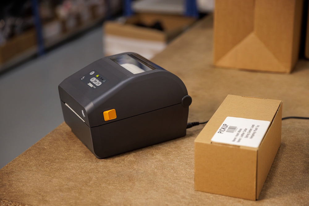 Zebra etikettskrivare på arbetsbänk bredvid kartong med nyutskriven etikett.